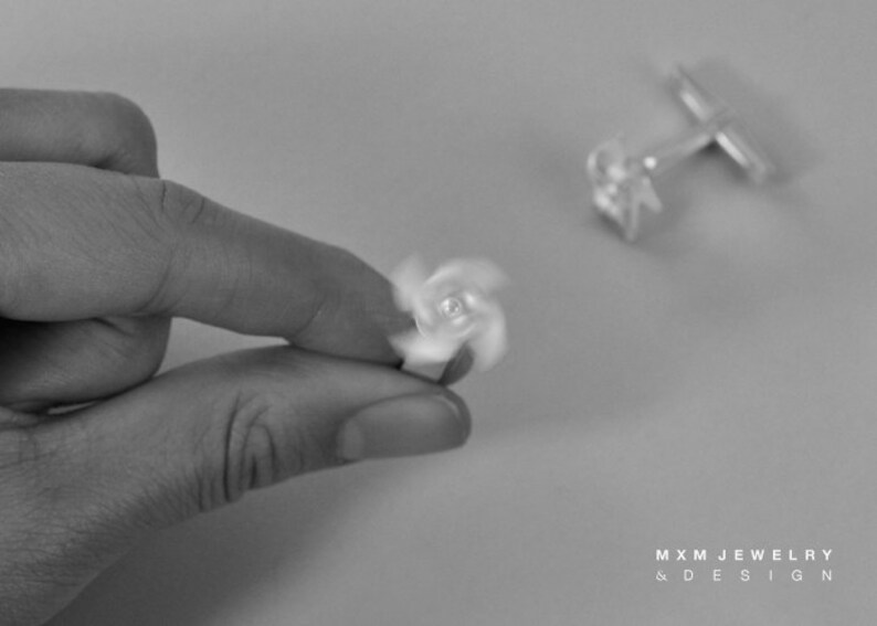 Puede incluir: Un par de gemelos de plata con un dise&ntilde;o giratorio. Los gemelos se muestran sobre una superficie blanca. El texto "MXM JEWELRY & DESIGN" es visible en la esquina inferior derecha de la imagen.