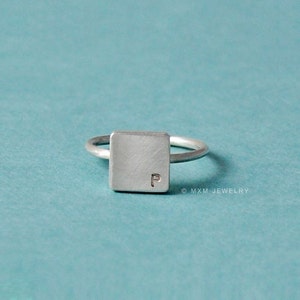 Puede incluir: Un anillo de plata con una banda de forma cuadrada que presenta la letra "P" grabada en la parte superior. El anillo est&aacute; sobre un fondo turquesa.