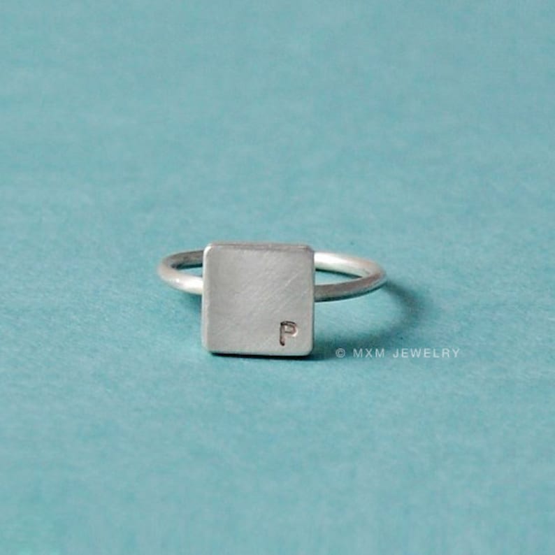 Puede incluir: Un anillo de plata con una banda de forma cuadrada que presenta la letra "P" grabada en la parte superior. El anillo est&aacute; sobre un fondo turquesa.