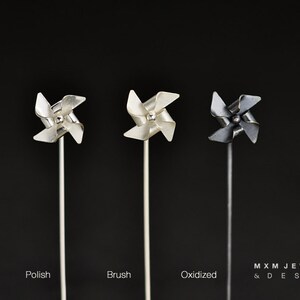 May include: Three silver pinwheel-shaped brooches in different finishes: polished, brushed, and oxidized. The brooches are displayed on a black background.