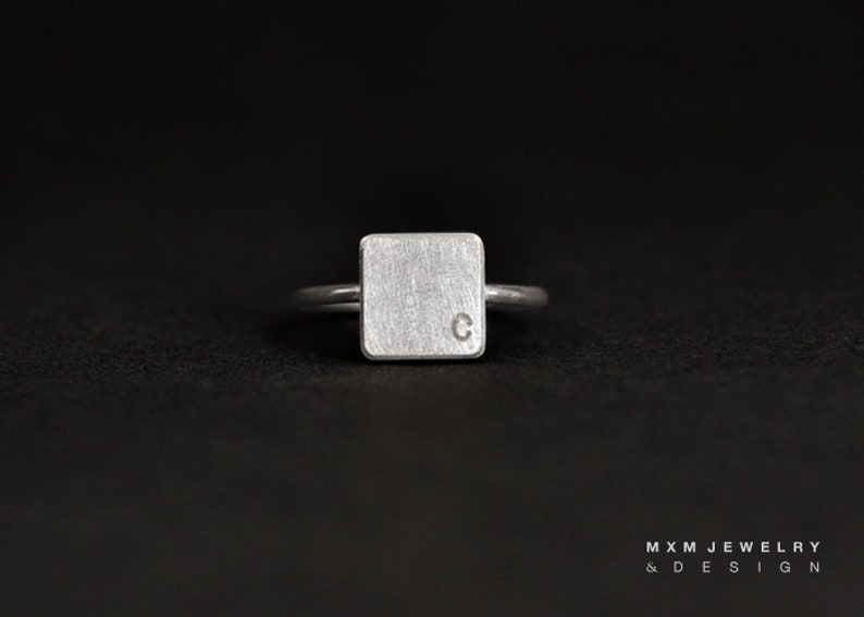 Puede incluir: Un anillo de plata con una banda cuadrada que presenta la letra "C" grabada en la parte superior.