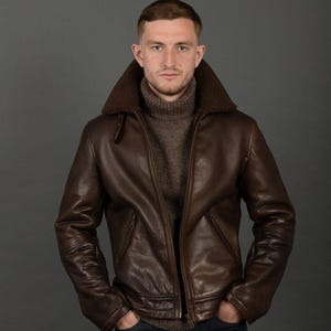 Puede incluir: Chaqueta de cuero marr&oacute;n con cuello de borrego y cremallera. La chaqueta tiene un dise&ntilde;o cl&aacute;sico con dobladillo y pu&ntilde;os acanalados. La persona lleva un jersey de cuello alto marr&oacute;n y vaqueros azules. El fondo es gris s&oacute;lido.