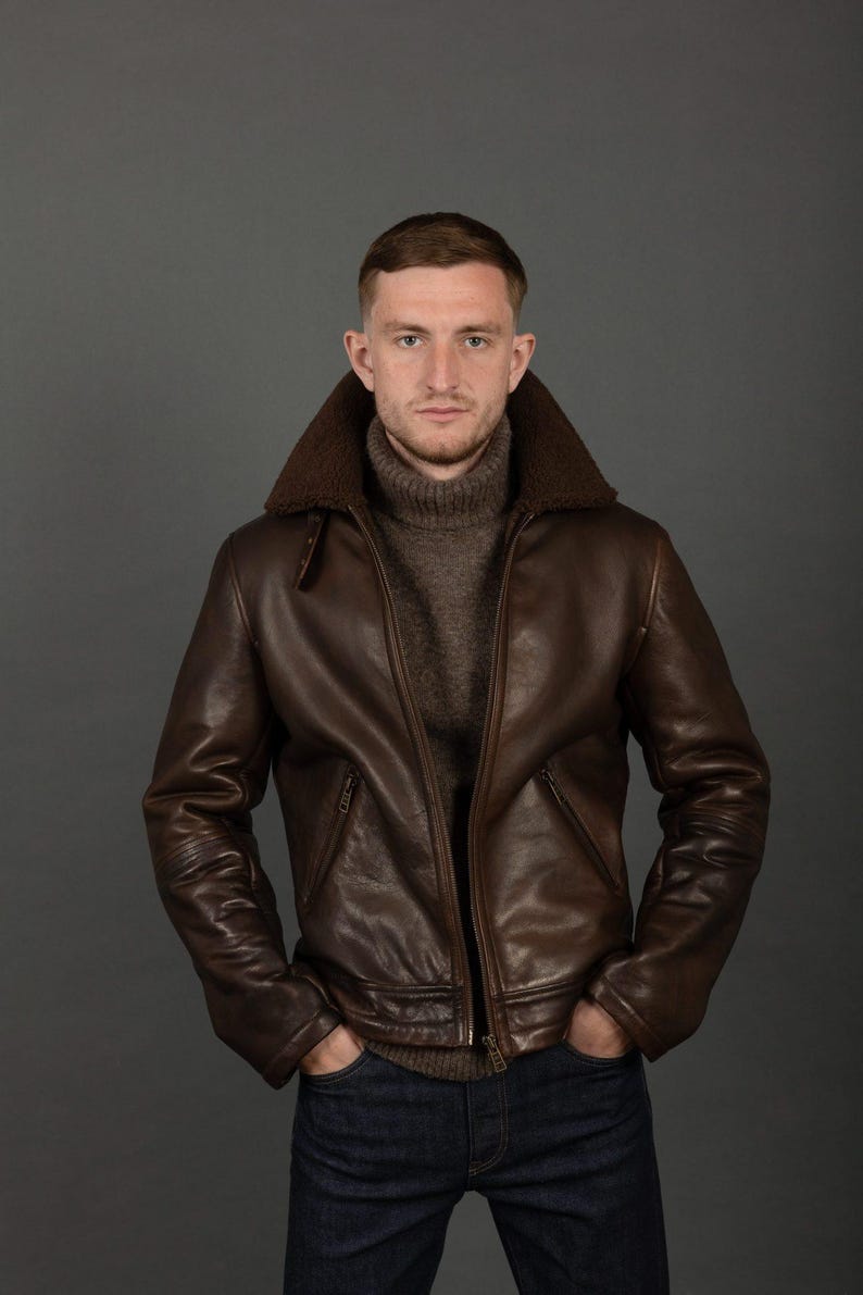 Puede incluir: Chaqueta de cuero marr&oacute;n con cuello de borrego y cremallera. La chaqueta tiene un dise&ntilde;o cl&aacute;sico con dobladillo y pu&ntilde;os acanalados. La persona lleva un jersey de cuello alto marr&oacute;n y vaqueros azules. El fondo es gris s&oacute;lido.