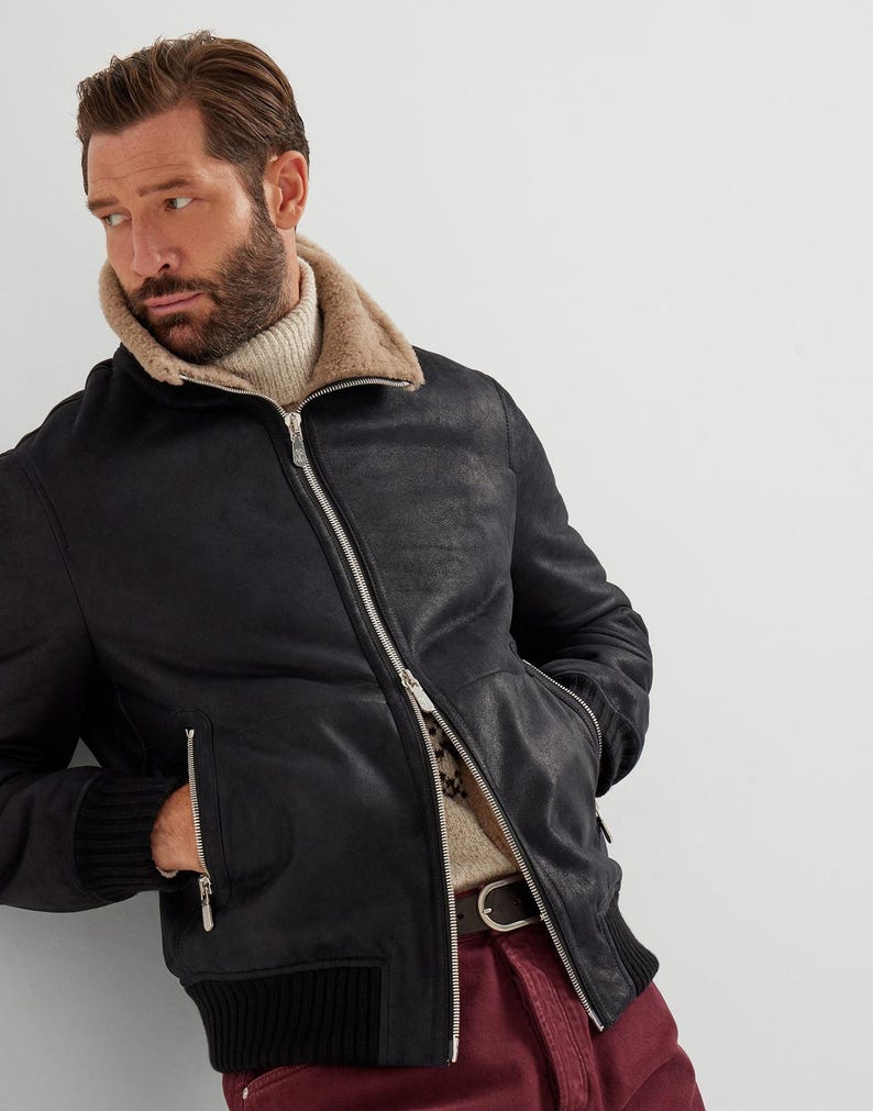 May include: Black leather bomber jacket with a shearling collar and a ribbed knit hem and cuffs. The jacket features a silver zipper closure and two side pockets. The model is wearing a cream-colored turtleneck sweater.