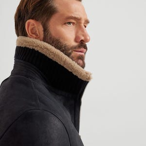 May include: A black leather jacket with a high, ribbed collar and a tan shearling lining. The jacket has a zipper closure and a textured surface. The model is wearing the jacket.
