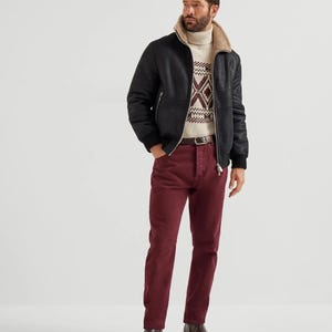 May include: A black leather jacket with a beige shearling collar and a patterned sweater. The person is wearing burgundy pants and brown boots. The jacket has a zipper closure and ribbed cuffs.