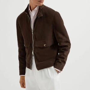 May include: A brown suede jacket with a zip-up front, two front pockets, and a stand-up collar. The jacket is worn over a white shirt and white pants. The jacket's design is simple and classic.