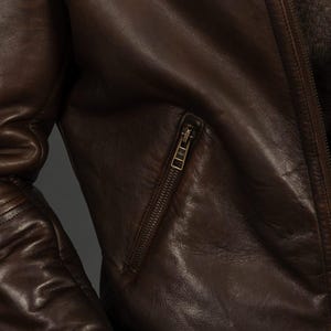 Puede incluir: Primer plano de una chaqueta de cuero marr&oacute;n con un bolsillo con cremallera y cierre de cremallera. La chaqueta tiene una superficie texturizada y un dise&ntilde;o cl&aacute;sico. El cuero parece desgastado, d&aacute;ndole un aspecto vintage.