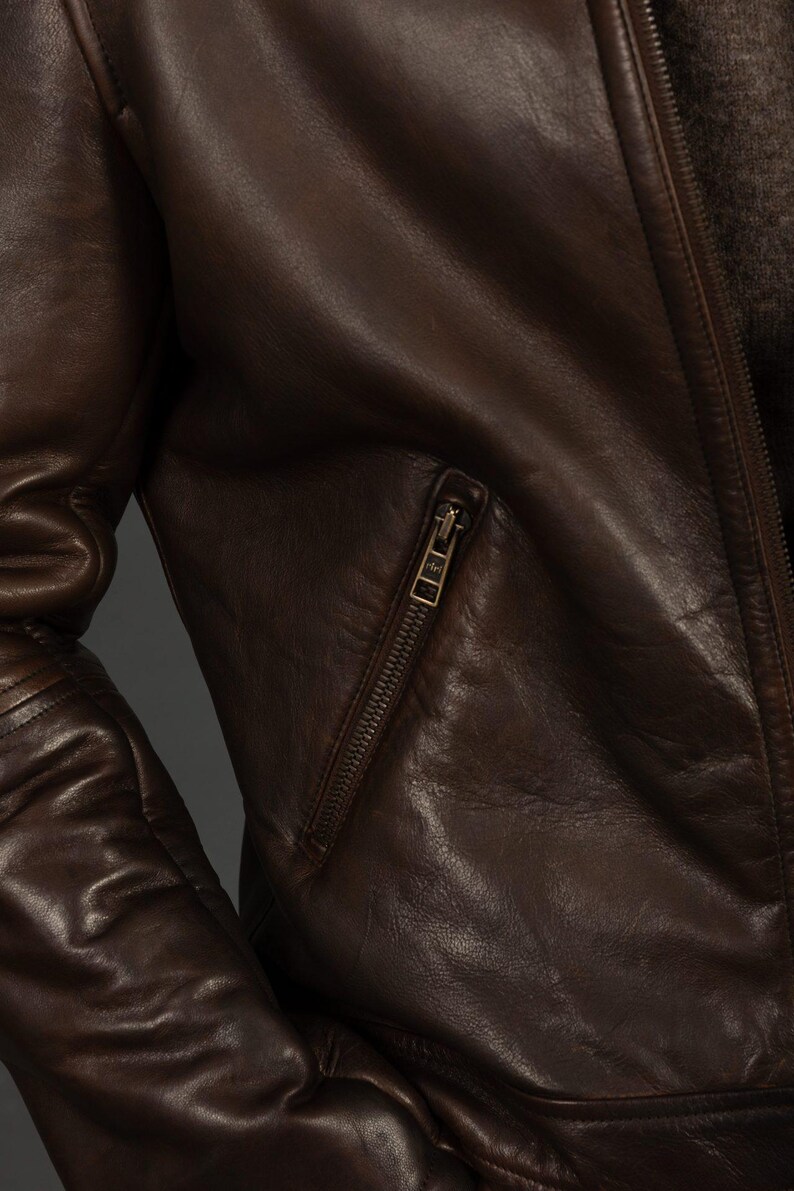 Puede incluir: Primer plano de una chaqueta de cuero marr&oacute;n con un bolsillo con cremallera y cierre de cremallera. La chaqueta tiene una superficie texturizada y un dise&ntilde;o cl&aacute;sico. El cuero parece desgastado, d&aacute;ndole un aspecto vintage.