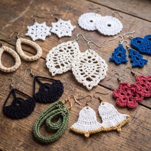 May include: A collection of various crochet earrings featuring diverse shapes including white snowflakes, beige teardrops, white leaves, blue hearts, black fans, red flowers, a green oval, and white bells with gold trim, all attached to metal earring hooks.