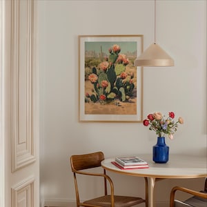 May include: A framed print of a desert landscape featuring a large cactus with pink flowers. The print is displayed on a white wall above a round table with a blue vase of flowers. A wooden chair is next to the table.