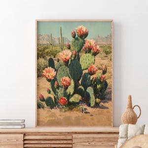 May include: A framed print of a desert landscape featuring a large prickly pear cactus with vibrant pink flowers. The cactus is set against a backdrop of mountains and other cacti, with a warm, sandy foreground.