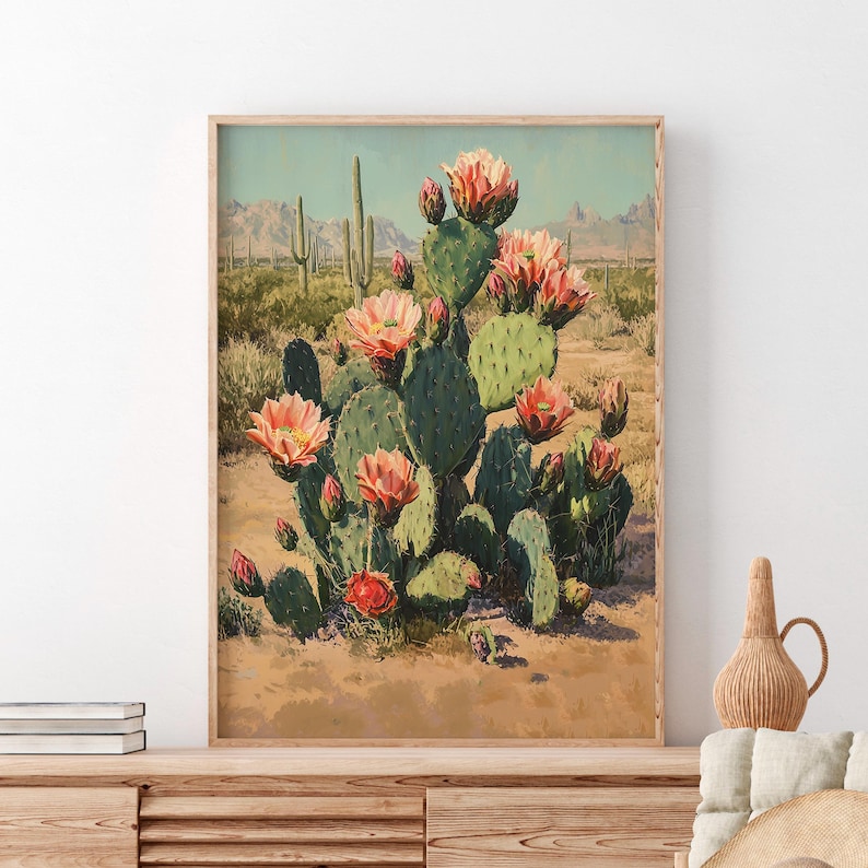 May include: A framed print of a desert landscape featuring a large prickly pear cactus with vibrant pink flowers. The cactus is set against a backdrop of mountains and other cacti, with a warm, sandy foreground.
