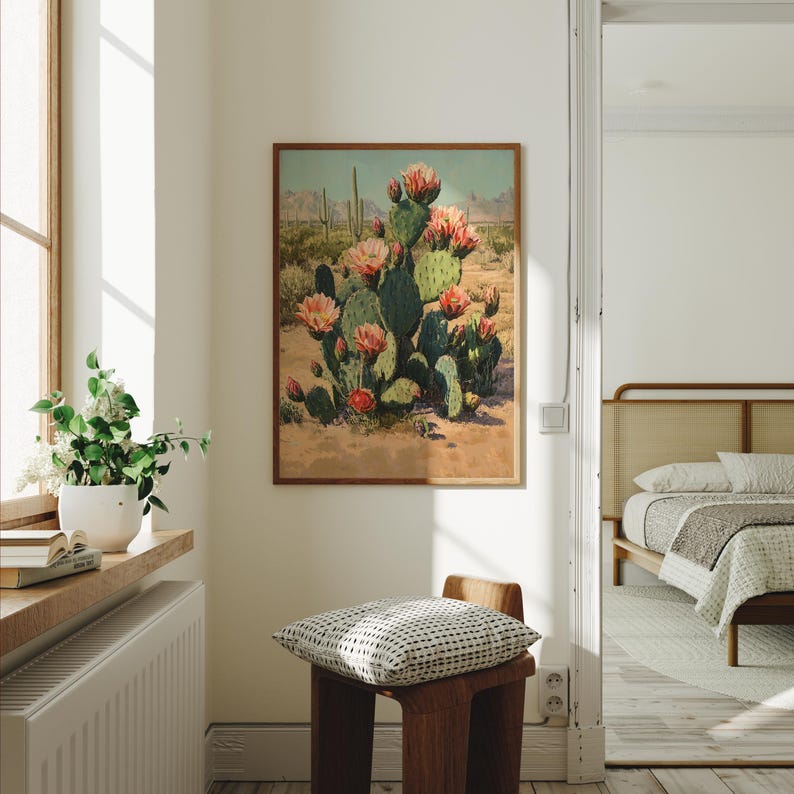 May include: A framed print of a desert landscape featuring a large cactus with pink flowers. The artwork is displayed in a room with a window, a wooden stool, and a glimpse of a bed. The color palette includes greens, pinks, and browns.