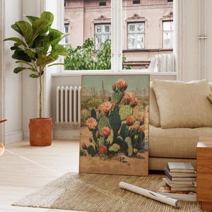 May include: A framed print of a desert landscape featuring a blooming cactus with pink flowers. The artwork is displayed indoors, leaning against a beige sofa, with a window and a potted plant in the background.