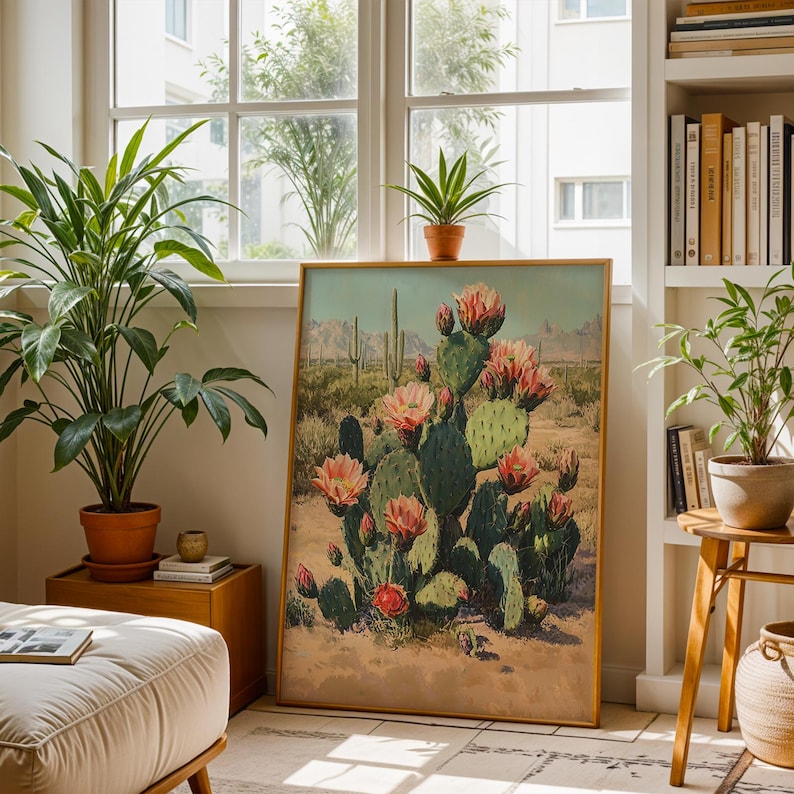 May include: A framed art print featuring a desert landscape with a blooming cactus. The artwork showcases a large green cactus with pink flowers, set against a backdrop of mountains and a sandy terrain. The frame is a warm, natural wood tone.
