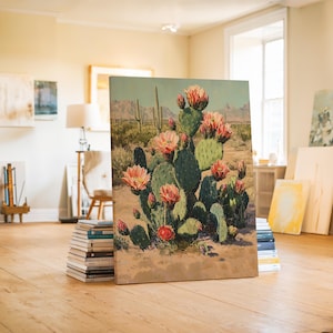 May include: A canvas print of a desert landscape featuring a blooming prickly pear cactus. The artwork showcases a vibrant green cactus with pink flowers, set against a backdrop of mountains and a sandy terrain. The painting is propped up on a stack of books.