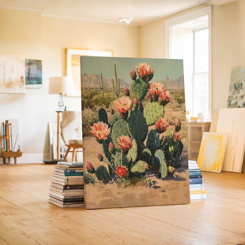 May include: A canvas print of a desert landscape featuring a blooming prickly pear cactus. The artwork showcases a vibrant green cactus with pink flowers, set against a backdrop of mountains and a sandy terrain. The painting is propped up on a stack of books.