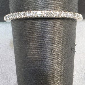 May include: A silver band ring with a row of small, sparkling gemstones. The ring is set against a dark, textured background, highlighting the brilliance of the stones. The ring appears to be a delicate piece of jewelry.