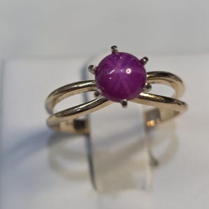 May include: A gold ring with a large, round, deep pink gemstone exhibiting a star effect. The ring has a double-band design, with the gemstone set atop the bands. The ring is displayed on a white surface.