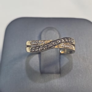 Puede incluir: Un anillo dorado con un diseño en espiral, engastado con pequeñas piedras brillantes. El anillo presenta dos bandas que se cruzan, creando un aspecto único y elegante. Las piedras están espaciadas uniformemente a lo largo de las bandas.