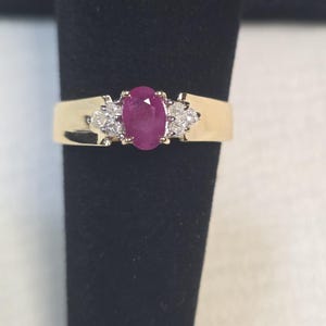Könnte beinhalten: Ein Goldring mit einem zentralen, oval geschliffenen, tiefrosa Rubin, der auf jeder Seite von drei kleinen, rund geschliffenen weißen Diamanten flankiert wird. Die Ringschiene ist glatt und poliert und zeigt ein klassisches Design.
