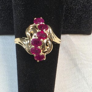 May include: A gold ring featuring a vertical row of five oval-cut vibrant pinkish-red gemstones set in a polished yellow gold band with swirling, organic metalwork accents that frame the central stones in a continuous, flowing design.