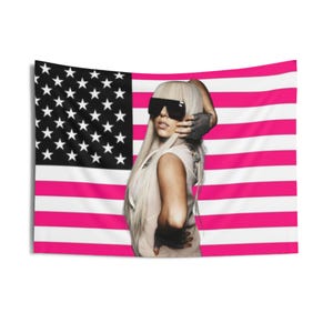 Puede incluir: Un tapiz de pared con una bandera estadounidense estilizada con rayas rosas y blancas y un campo de estrellas en blanco y negro. Una persona con cabello rubio largo y gafas de sol está colocada frente a la bandera.