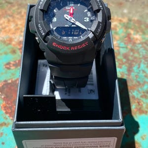 May include: A black Casio G-Shock watch with a round analog-digital face, red accents, and white hour markers. The watch features the text G-SHOCK and SHOCK RESIST on the bezel and is presented in a black box with a battery safety warning label.