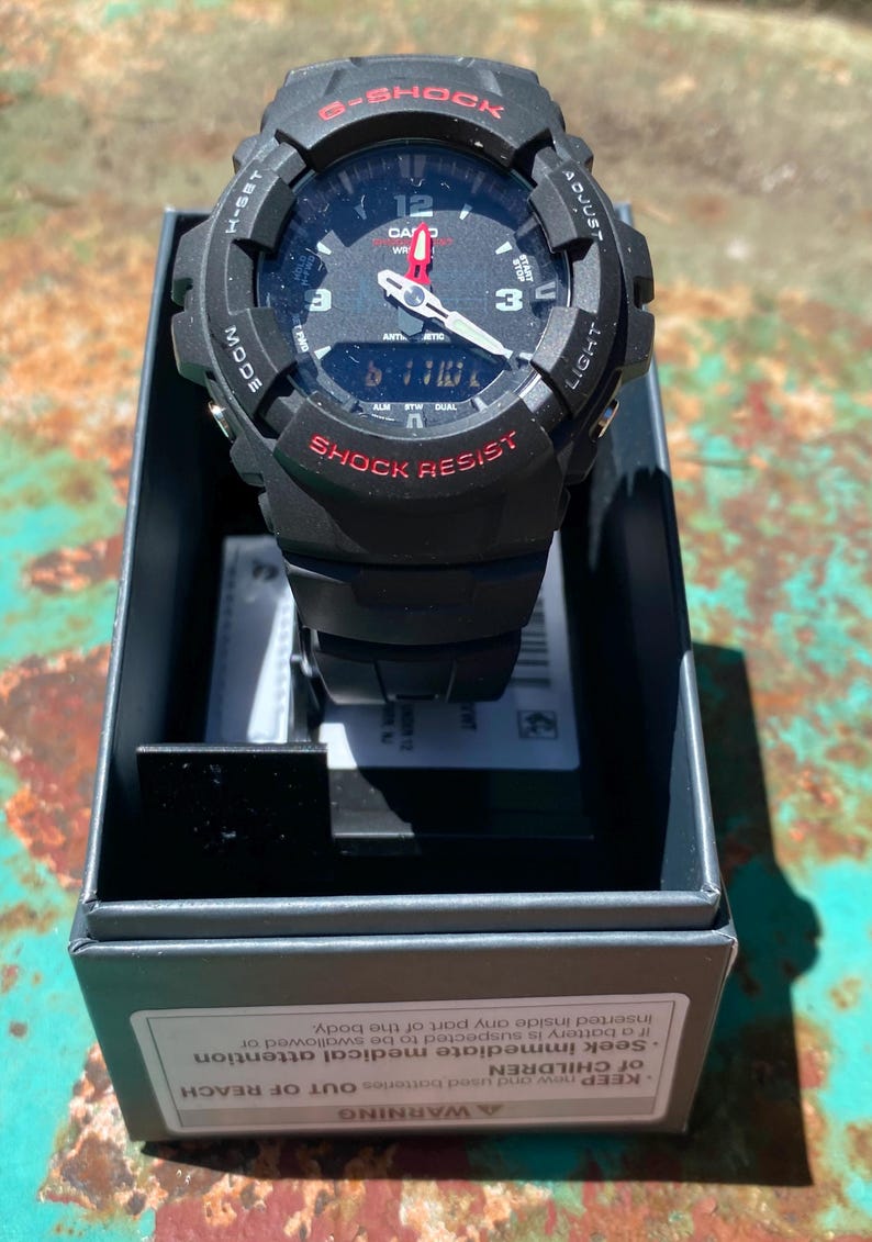 May include: A black Casio G-Shock watch with a round analog-digital face, red accents, and white hour markers. The watch features the text G-SHOCK and SHOCK RESIST on the bezel and is presented in a black box with a battery safety warning label.