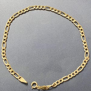 May include: A gold-colored Figaro chain bracelet. The bracelet features a pattern of three small circular links followed by one elongated oval link. The clasp is a lobster claw style. The bracelet is displayed on a dark gray surface.
