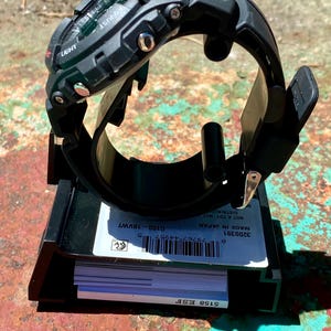 May include: A black G-Shock digital watch with a resin strap and metallic side buttons, mounted on a black display stand. The base features a white label with a barcode, the text G100-18VWT, 3200391, MADE IN JAPAN, and 5158 ESF.