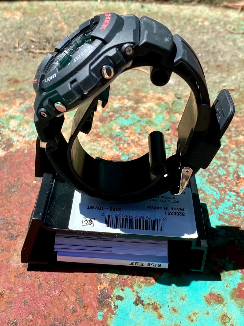 May include: A black G-Shock digital watch with a resin strap and metallic side buttons, mounted on a black display stand. The base features a white label with a barcode, the text G100-18VWT, 3200391, MADE IN JAPAN, and 5158 ESF.