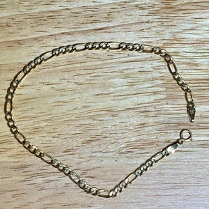 May include: A gold-colored Figaro chain bracelet. The bracelet features a pattern of three small round links followed by one elongated oval link. The clasp is visible at the end of the bracelet. The bracelet is displayed on a wooden surface.
