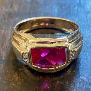 May include: A gold ring with a large, rectangular, deep red gemstone. The ring has a wide band with ridged detailing and small accent stones on either side of the gemstone. The ring is set on a wooden surface.