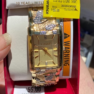May include: A gold-tone rectangular wristwatch with a gold dial and hour markers. The watch band features textured gold links accented with clear stones. The watch is housed in a red box with a white display cushion and includes a yellow clearance sticker.