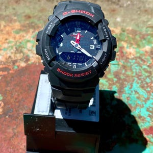 May include: A black G-Shock watch with a round analog-digital face, featuring red text accents, white hour and minute hands with a red second hand, and a black resin strap. The bezel is marked with H-SET, ADJUST, MODE, and LIGHT functions.
