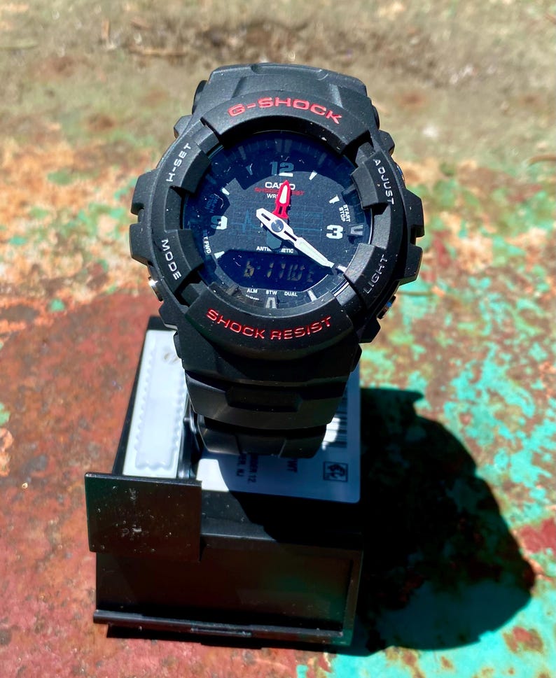 May include: A black G-Shock watch with a round analog-digital face, featuring red text accents, white hour and minute hands with a red second hand, and a black resin strap. The bezel is marked with H-SET, ADJUST, MODE, and LIGHT functions.