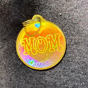 May include: A gold-colored metal keychain with a round tag that says "MOM" in a colorful, swirling font. The tag is surrounded by a decorative border of hearts and swirls.
