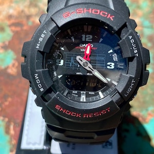 May include: A black G-Shock watch with a round analog-digital face, red lettering, and white hour markers. The dial features a grid pattern, a red second hand, and silver hour and minute hands. The bezel is marked with H-SET, ADJUST, MODE, and LIGHT.