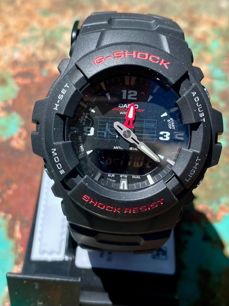 May include: A black G-Shock watch with a round analog-digital face, red lettering, and white hour markers. The dial features a grid pattern, a red second hand, and silver hour and minute hands. The bezel is marked with H-SET, ADJUST, MODE, and LIGHT.