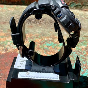 May include: A black G-Shock digital watch mounted on a black plastic display stand. The watch features a rugged, textured bezel with visible side buttons labeled H-SET and MODE. A white label on the base includes the text MADE IN JAPAN.