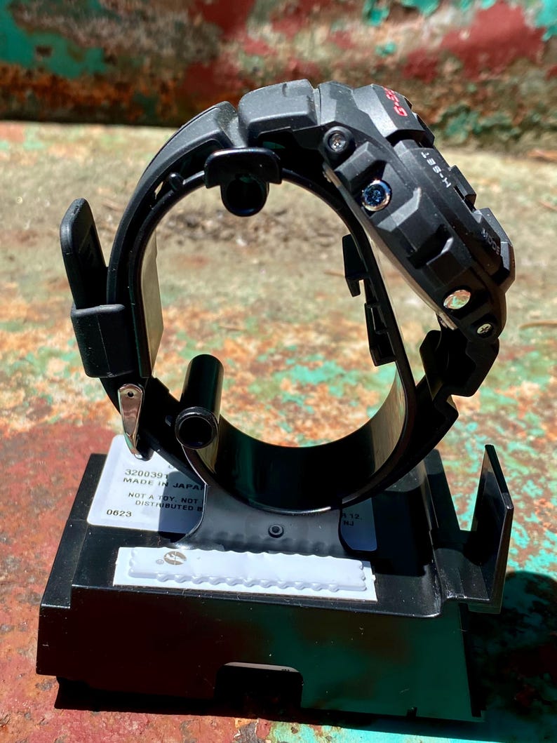 May include: A black G-Shock digital watch mounted on a black plastic display stand. The watch features a rugged, textured bezel with visible side buttons labeled H-SET and MODE. A white label on the base includes the text MADE IN JAPAN.