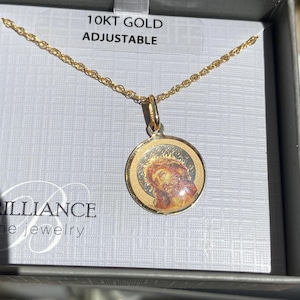 Op de afbeelding: Een goudkleurige ketting met een ronde hanger met een afbeelding van Jezus. De hanger is gezet in een gouden frame en hangt aan een fijne gouden ketting. De verpakking leest "10KT GOLD ADJUSTABLE".