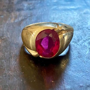 May include: A gold ring featuring a large, oval, deep red gemstone. The ring has a thick, rounded band with a polished finish. The gemstone is the focal point, set centrally within the ring.