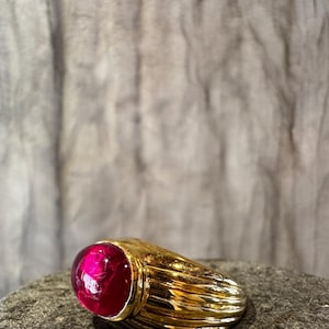 May include: A gold ring with a large, oval-shaped, cabochon ruby gemstone. The ring has a textured band with vertical grooves.
