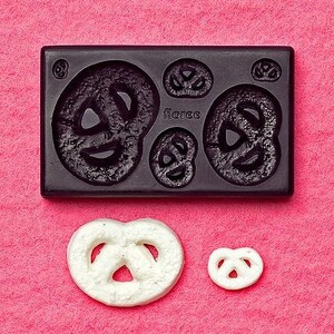 Puede incluir: Un molde de silicona negro con formas de pretzel y dos pretzels blancos sobre un fondo rosa. El molde tiene la palabra "floree". Los pretzels son de diferentes tamaños, uno grande y uno pequeño.