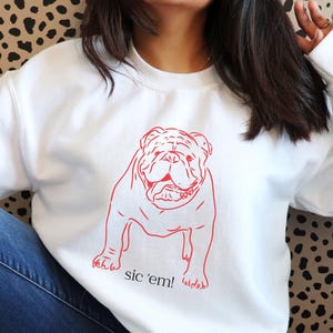 May include: White sweatshirt featuring a red outline of a bulldog with the text "sic 'em!". The sweatshirt is worn with blue jeans. The background has a black and brown spotted pattern.