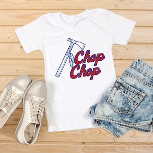 May include: White t-shirt with a blue and red graphic of a tomahawk and the words "Chop Chop". The shirt is paired with denim shorts and silver sneakers, all arranged on a wooden surface.