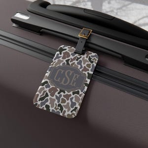 May include: A luggage tag with a camouflage pattern in shades of brown, gray, and white. The tag features a dark gray rectangular emblem with the initials "CSE" in a stylized font. The tag is attached to a black leather strap with a gold-colored buckle.